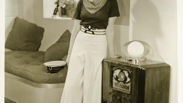 A woman wearing white trousers and a black top stands  leaning against an old radiogram.