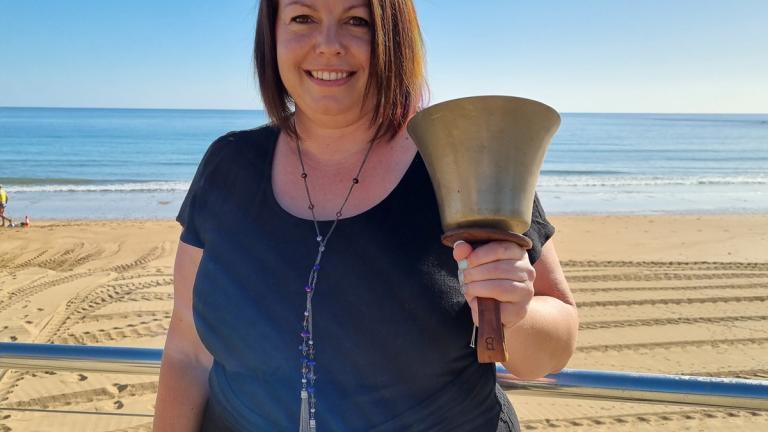 Woman holds a large bell. She is standing outdoors with a sandy beach and ocean behind her.
