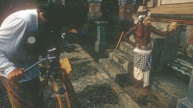 A camera man filming a balinese man in cultural dress and mask