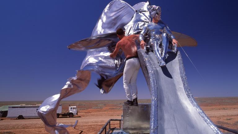 Guy Pearce sitting on a giant silver stilleto shoe on top of the Priscilla bus.