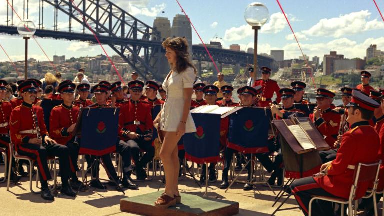 A woman in a white minidress stands, surrounded by a military band. The Sydney Harbour Bridge is in the background.