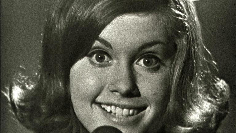 Close up of Olivia Newton-John smiling and looking directly at the camera with a microphone in front of her face, partially covering her chin, from an episode of 'Boomeride' when she was aged 16.