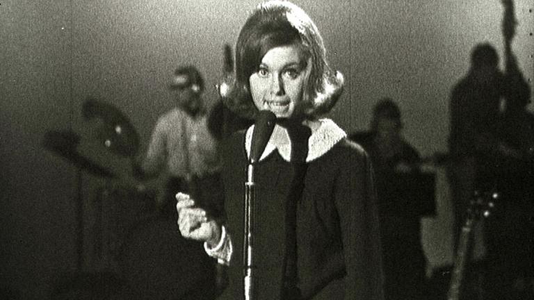 Olivia Newton-John at age 16 in a TV studio standing behind a microphone singing.