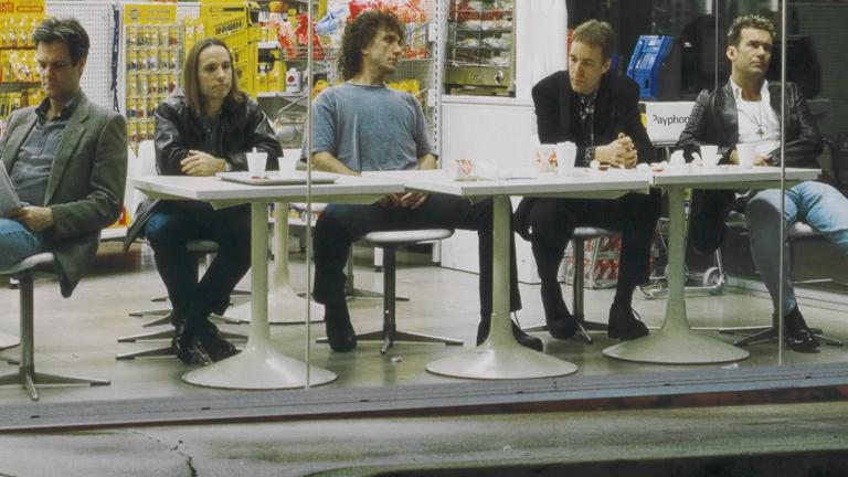 The five members of the band Cold Chisel seated at white plastic tables at a service station. From left to right: Don Walker, Phil Small, Ian Moss, Steve Prestwich and Jimmy Barnes.