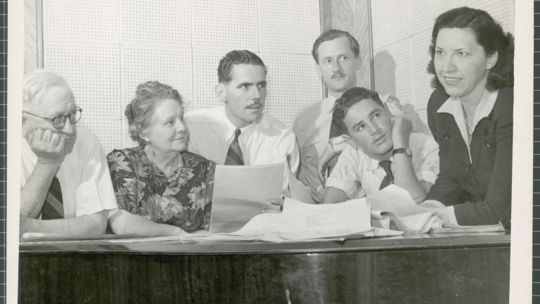 Grace Gibson (far right) leans on a desk. Next to her are a group of people all sitting at the desk and looking up at her.