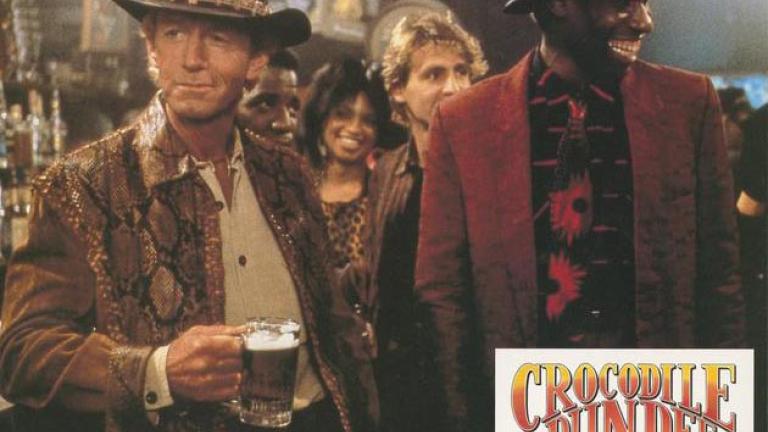 Lobby card depicting Mick Dundee standing in a bar with a beer in his hand and a guy smiling next to him like Mick's just said something funny
