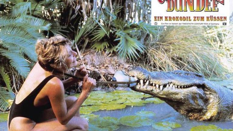 German lobby card for Crocodile Dundee depicting Sue Charlton wrestling to get a water bottle off from around her neck because a crocodile has got a hold of it 