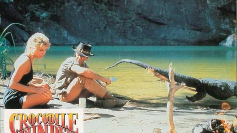Lobby card depicting Linda Kozlowski as Sue Charlton and Paul Hogan's Mick 'Crocodile’ Dundee in the outback with a crocodile roasting on a spit made of tree branches
