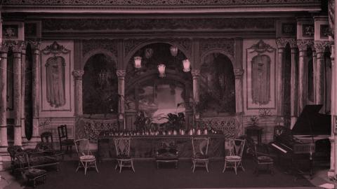 A tinted image of an early 20th century stage before a musical performance. There are a row of chairs for the performers, a piano and a table with a row of handbells.