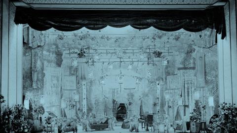A tinted image of an early 20th century stage before a Corricks family musical performance. It features a piano, an array of chairs and armchairs for the performers, and decorative flowers both on the stage and suspended above it.