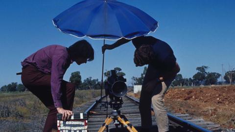 A man holds an umbrella over a film camera positioned on railway tracks while another man holds a clapperboard