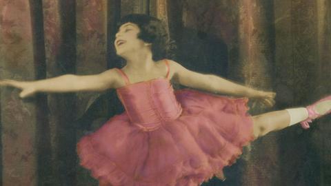 A hand-coloured still of Annette Kellerman as a child. She is dressed in a pink ballet tutu with ballet slippers and is striking a pose with her arms and on leg out.