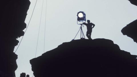 A member of a film crew stands silhouetted next to a standing light on an outcrop of rock