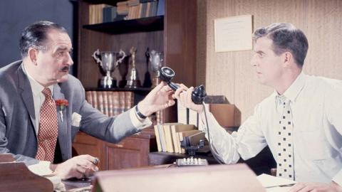 Two men in business attire are seated at an office desk. One is handing the telephone receiver to the other.