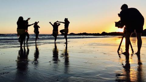 A camera filming silhouetted Aboriginal dancers on a Byron Bay beach at sunrise