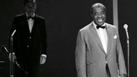 Jazz musician and singer Louis Armstrong standing in front of a microphone in a TV studio, smiling. Another musician is pictured beside him holding an instrument. Circa 1964.