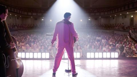 Actor Austin Butler in a scene from the film ELVIS. He is pictured from behind wearing a bright pink suit and standing on a stage in front of a large crowd in a theatre. 