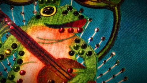 Detail of a costume from the film ELVIS showing a green frog playing a guitar. The diamante fabric of the costume is accented with sparkling jewels.