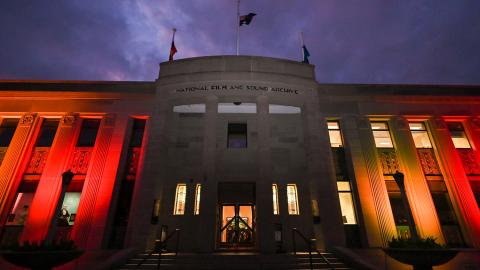 Front facade of an art deco style building with columns at the front and a sign saying National Film and Sound Archive. The building is lit up with red and orange lighting at night time.