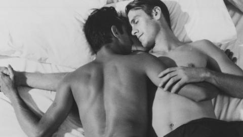 Two young men lying on a bed in an embrace. They are both shirtless and one is lying face down and the other facing up, with their faces turned toward eachother.