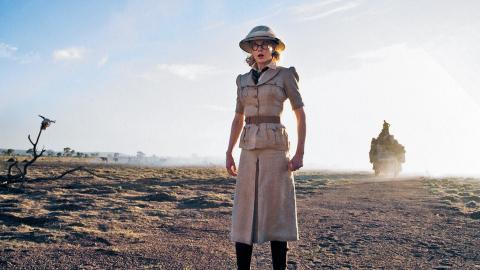 Full-length, wide shot of actor Nicole Kidman in a scene from the film Australia. She's standing in the dusty outback wearing a safari-style outfit of a beige skirt, belted shirt and hat with long dark brown boots. 