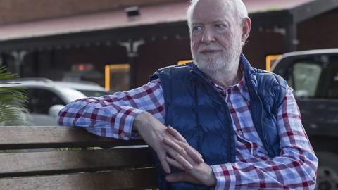 Film critic and author David Stratton seated on a bench outdoors. He is wearing a check shirt and vest and looking off to his right. 