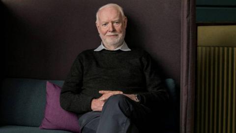 Film critic and author David Stratton seated on a sofa looking directly at the camera.