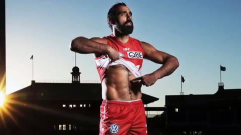 AFL football player Adam Goodes in the middle of a sports ground. He is wearing his Sydney Swans red and white uniform and lifting up the front of his shirt to point to the colour of his skin. 