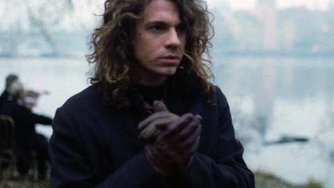 Head and shoulders shot of singer Michael Hutchence wearing a coat and gloves. There is a river and two people in the background.