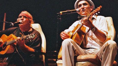 Kev Carmody and Paul Kelly on a stage seated in front of microphones and playing their guitars.