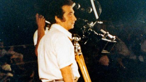 Filmmaker Giorgio Mangiamele standing outside in the dark next to a movie camera on a tripod. 