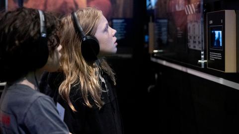 Two kids wearing headphones and looking at writing and a screen on the wall of an exhibition.