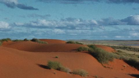 Orange-red sand-dunes of Southern Arrernte country, dotted with green scrub and with low-level clouds in a blue sky