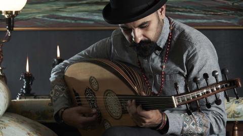 Joseph Tawadros wearing a black bowler hat and seated on a divan, playing an oud