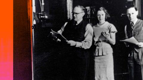 A man and two women in a recording studio standing next to a big radio microphone. They are holding scripts in their hands.