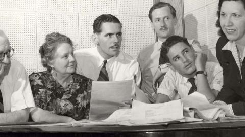 1940s radio series producer Grace Gibson leans on a table with pages of a script while four white men in plain shirts and ties and a woman wearing a floral-patterned dress look on