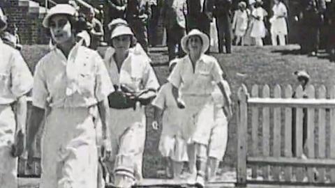 A team of women cricketers from the 1930s walking onto a cricket field in white uniforms.
