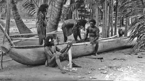 A group of Torres Strait Islander men building a canoe by the water, surrounded by palm trees in the early 1920s.
