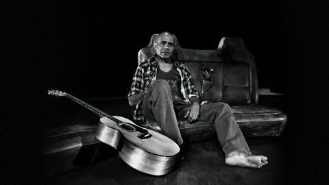Archie Roach rests against an old car seat in a studio with his guitar resting nearby.