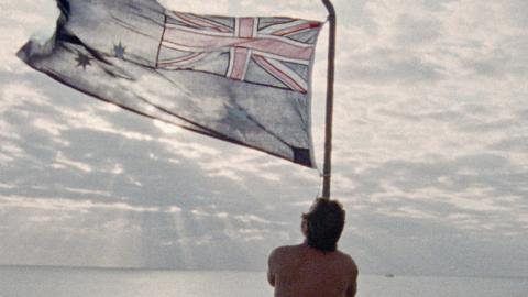 Man waving an Australian flag, set against a pale blue, cloudy sky.