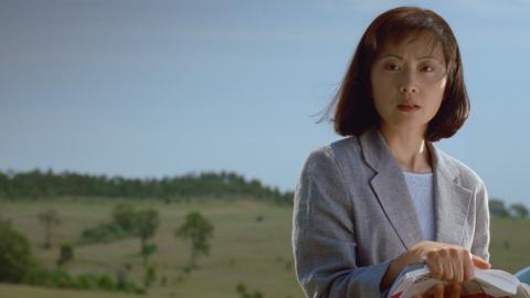 A young Asian woman dressed in a suit jacket pictured from the waist up in front of a green field and hills with trees in the background