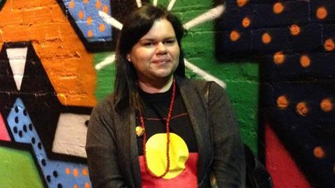 Tasha James standing in front of a colourful wall and wearing shirt bearing the Aboriginal flag