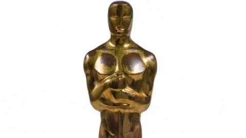 A gold Academy Award statue seen from the waist up, with a man clutching the handle of a sword