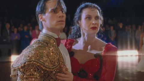A young man and woman pictured from waist up on a dance floor wearing ballroom costumes look anxiously at something off camera