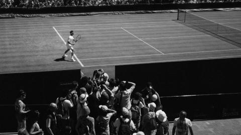 A male tennis player on the court at Wimbledon, watched by a scrum of press photographers