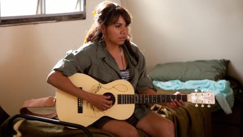 A woman sitting on a bed playing an acoustic guitar.