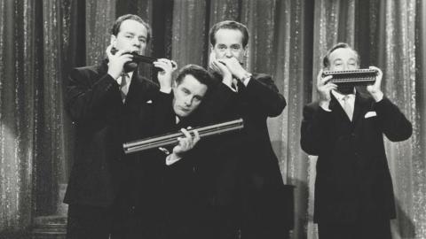 The members of the Horrie Dargie Quintet pose in front of a curtain, each member holding a harmonica