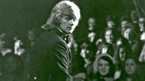 John Farnham sitting on stage in front of an audience, looking back over his right shoulder at the camer.