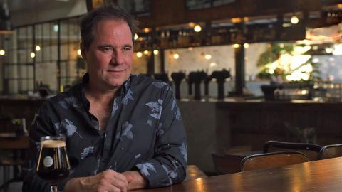 Waist-up shot of actor Paul Mercurio sitting at a restaurant table with a bar in the background. 