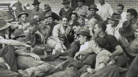 Actor Charles Farrell reclines on the ground surrounded by a crowd of men.